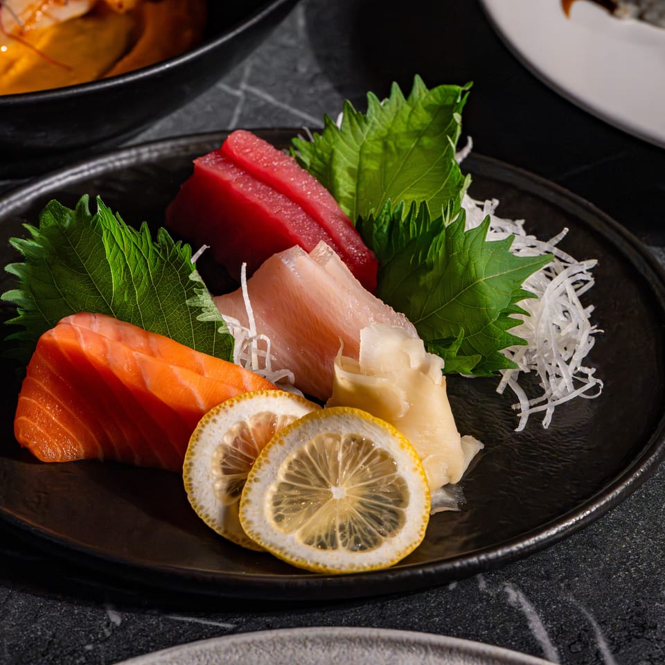 Japanese sashimi platter with salmon, tuna, white fish, shiso leaves, shredded daikon, lemon slices, and ginger on a black plate.
