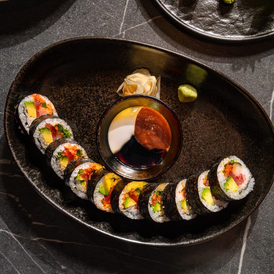 Sushi rolls with avocado, salmon, and roe arranged on a black plate beside dipping sauce, pickled ginger, and wasabi.