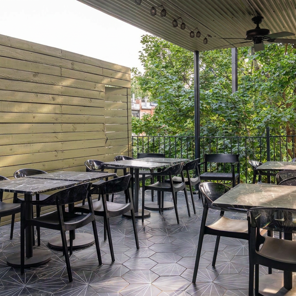 Covered restaurant patio with black tables and chairs, wooden privacy wall, ceiling fan, and leafy trees beyond the railing.